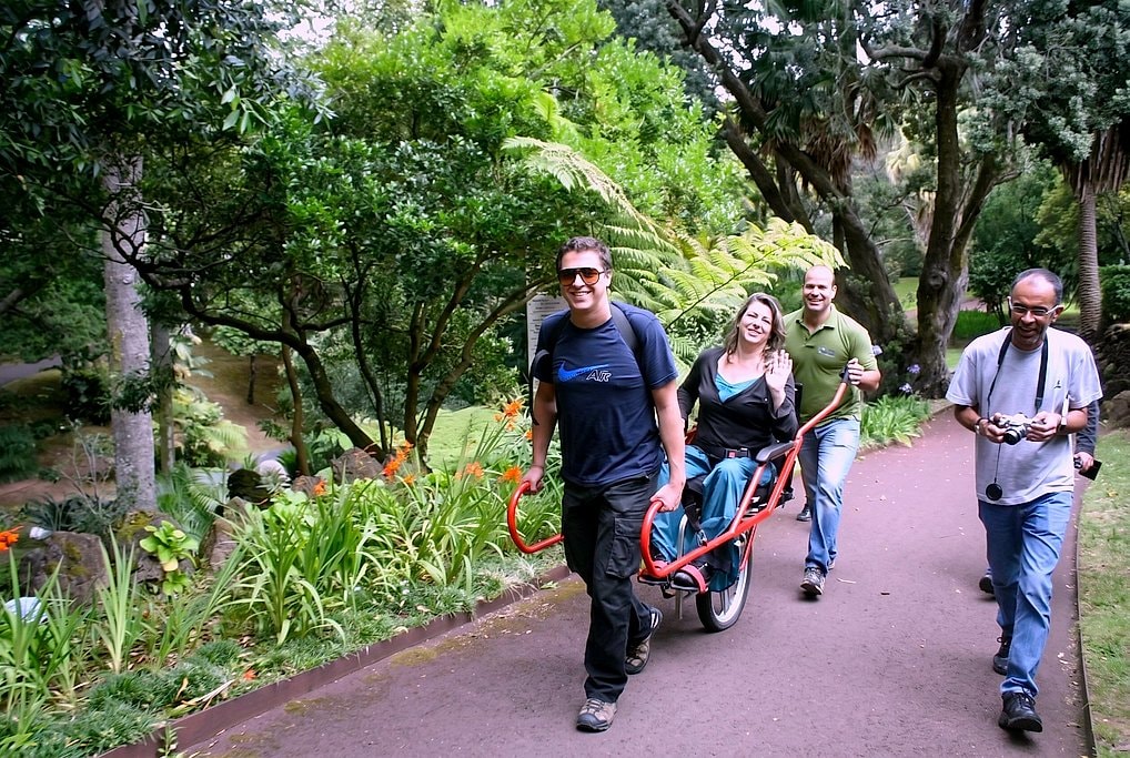 Two monitors guide a smiling seated rider in the Joelette chair along a garden path with orange tropical flowers.