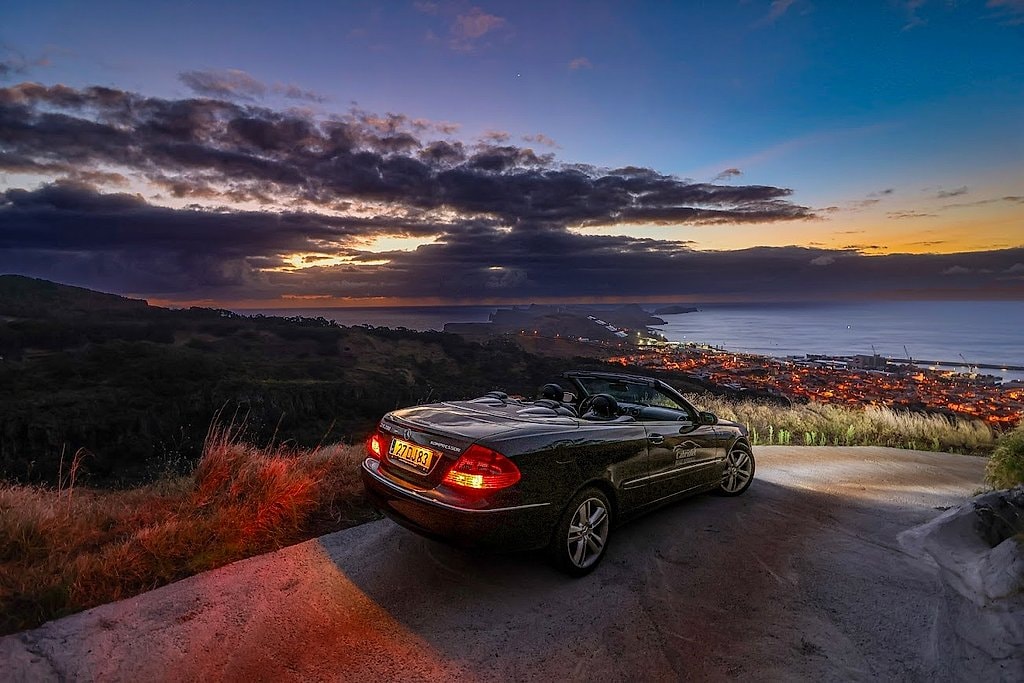 Convertible parked on a hilltop road at dusk, overlooking a lit-up Madeira coastal town.