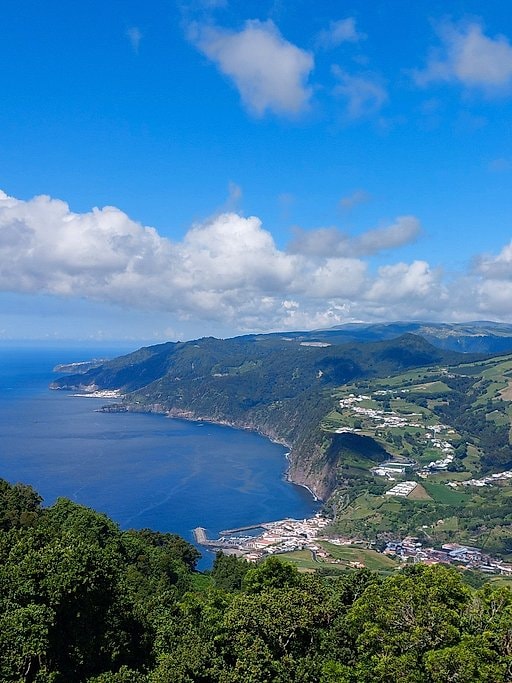 Aerial view of rugged green cliffs curving around a deep blue bay with a village at the shoreline