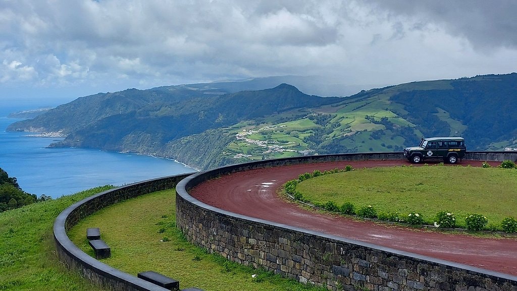 4x4 vehicle parked at a red-paved lookout platform overlooking layered green hills and the coastline