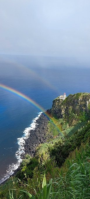 A rainbow arcs over ocean cliffs with a lighthouse on a rocky headland after passing rain