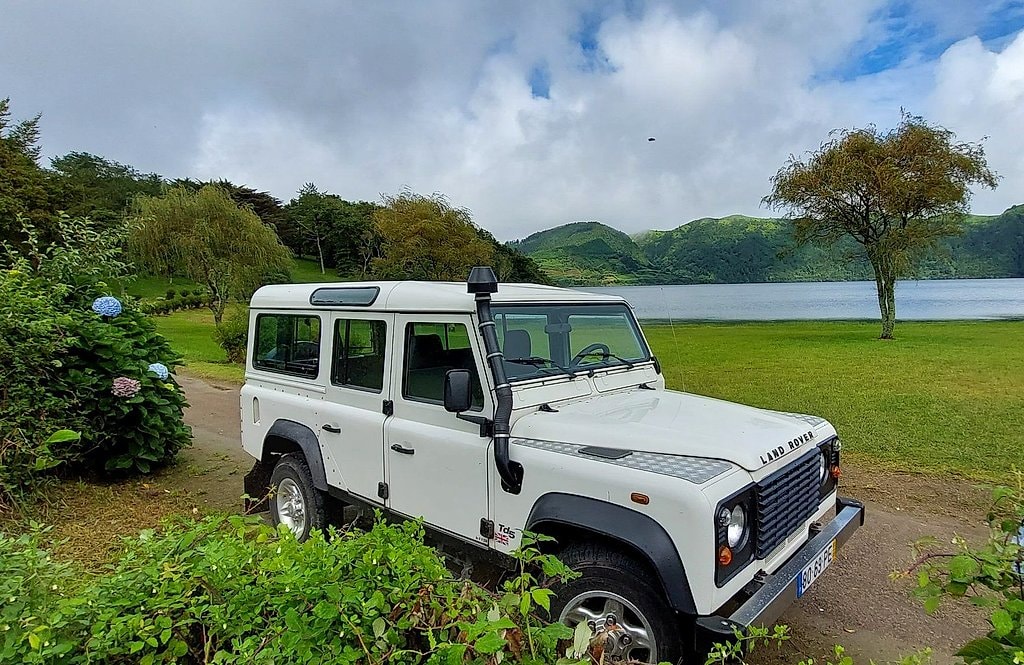 White 4x4 jeep parked by a green volcanic lake, with hydrangeas and misty hills in the background