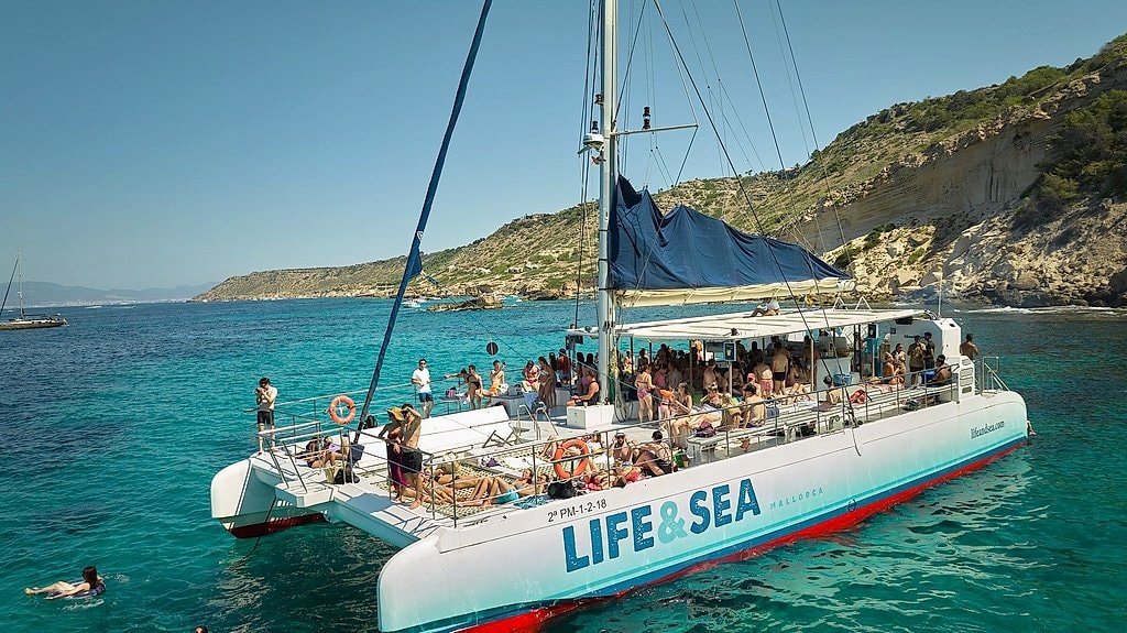 Catamaran deck crowded with passengers anchored near steep cliffs and a swimmer nearby.