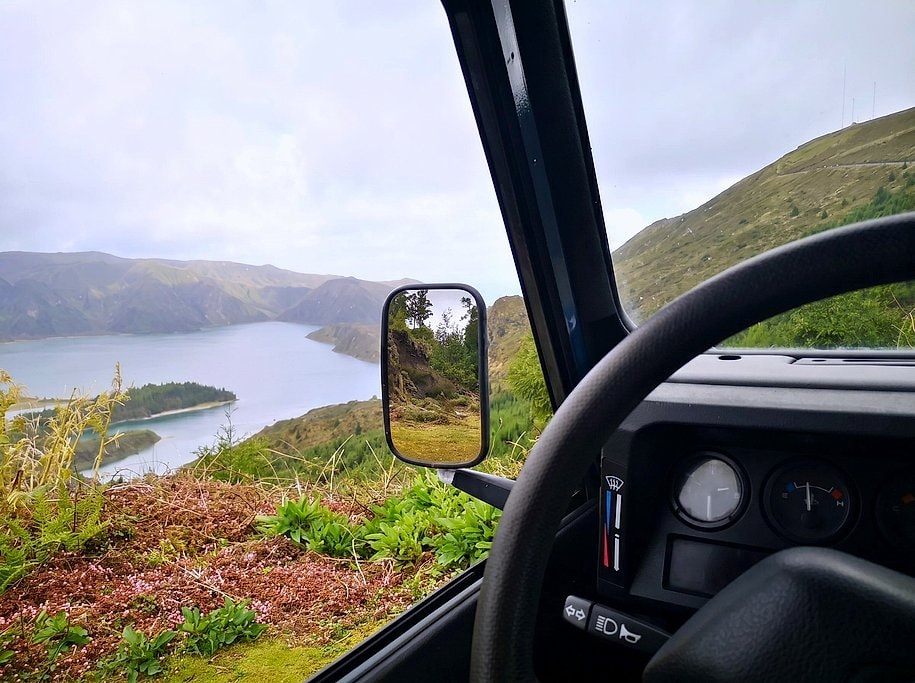 View through the jeep's side mirror over a turquoise lake and green valley on the mountain route