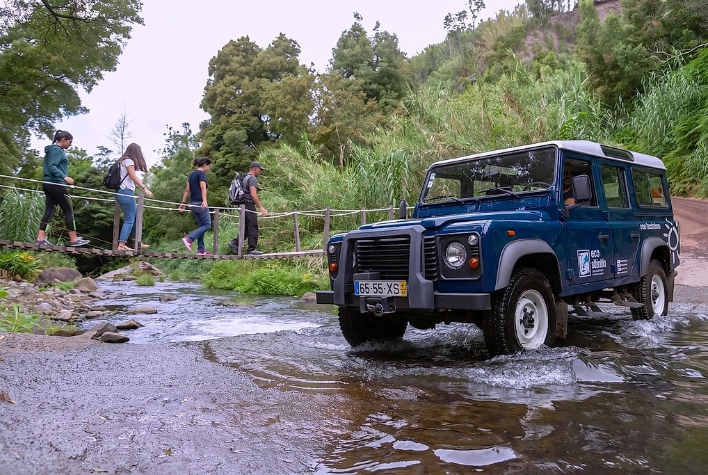 Jeep fording a shallow river while hikers cross a wooden footbridge alongside