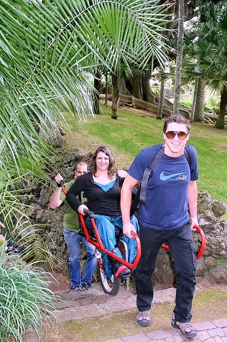 Close view of the red Joelette chair carried over stone steps between palm fronds on a garden trail.