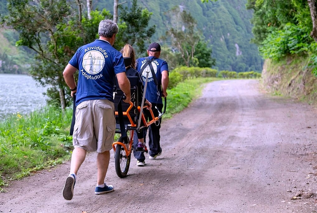 Two guides walk the orange Joelette chair along a dirt trail beside Sete Cidades lake, forested slopes behind.