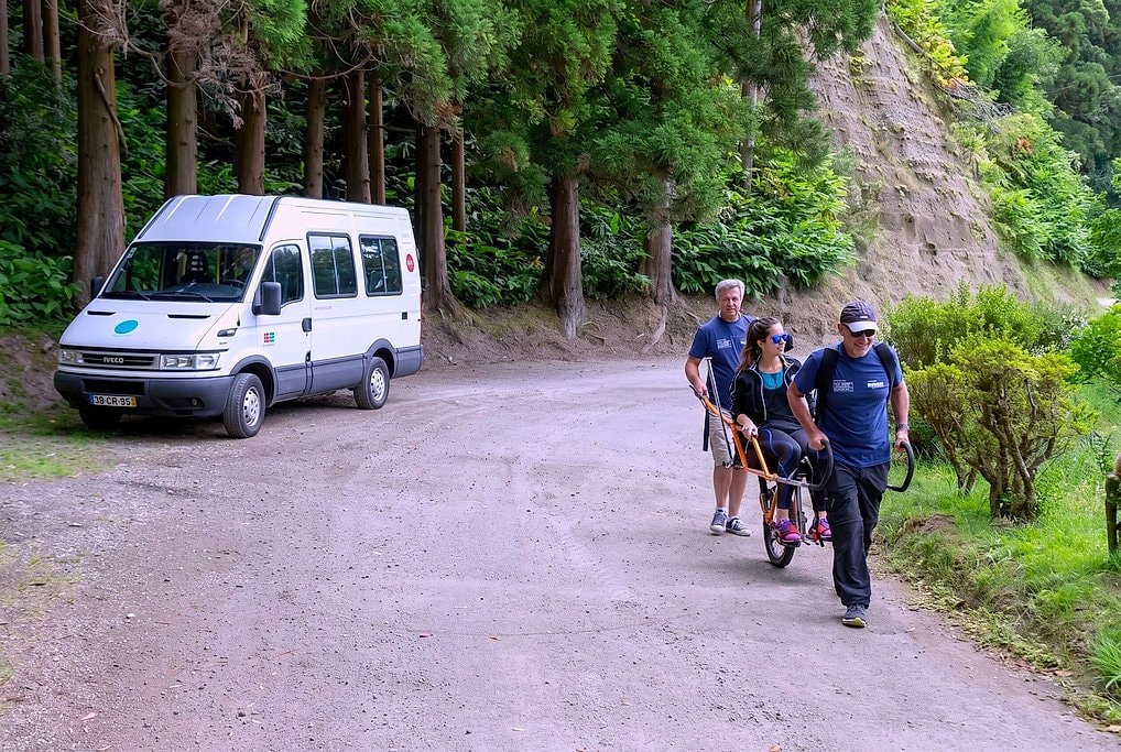 The orange Joelette chair with seated rider nears a parked support van on a forest dirt road.