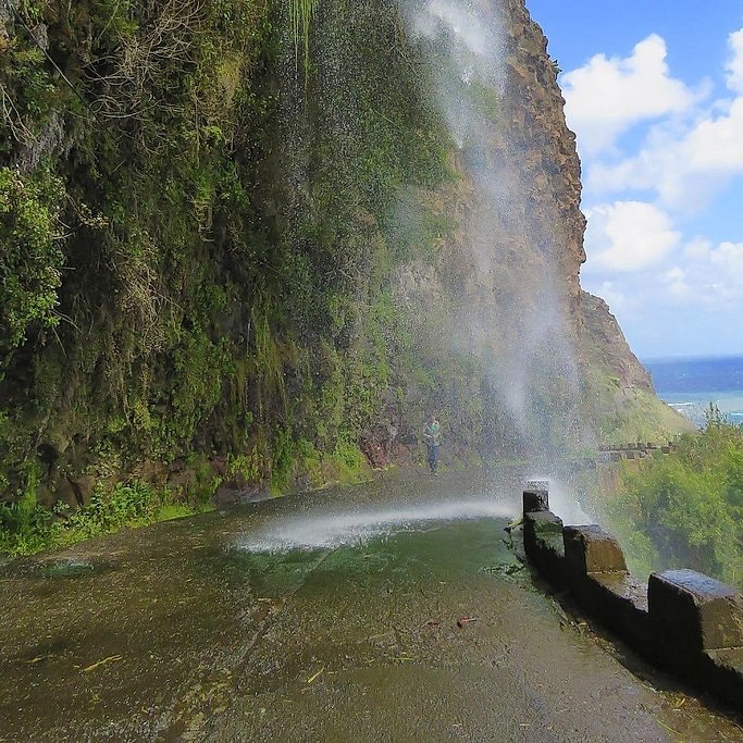 Cascata dos Anjos waterfall spraying across a cliff road, with someone standing beneath the falls.