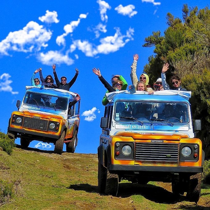 Two open-top jeeps full of cheering tourists driving along a grassy hillside in Madeira.