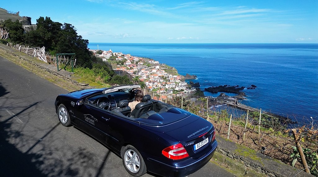 Black cabriolet convertible parked on a hillside road above a Madeira coastal town.