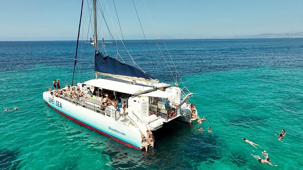 Catamaran anchored in turquoise water with passengers relaxing on deck and swimmers nearby.