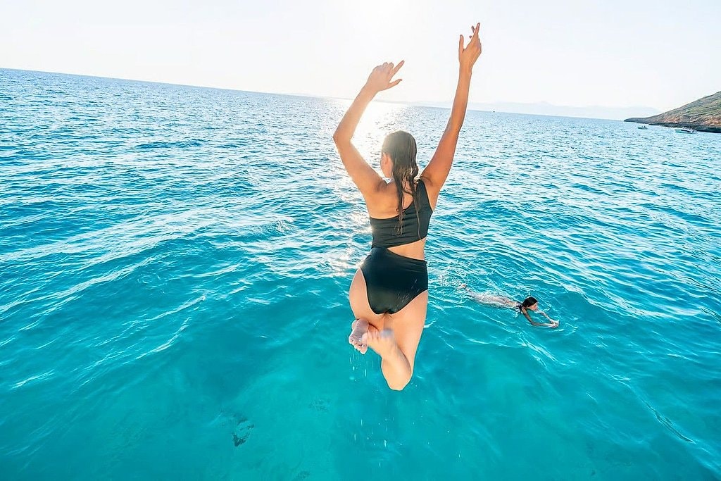 Woman leaping off the boat into clear water with arms raised overhead.