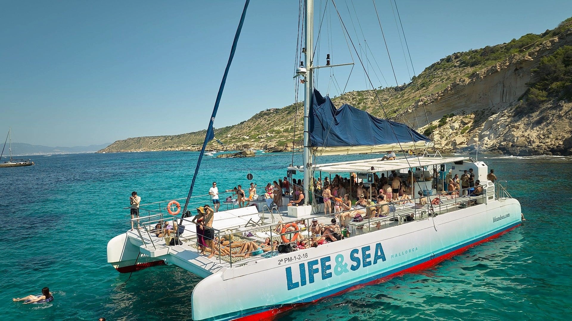 Catamaran deck crowded with passengers anchored near steep cliffs and a swimmer nearby.