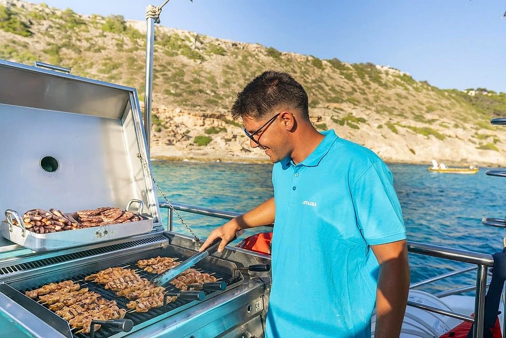 Crew member grilling meat and sausages on the boat's barbecue with cliffs behind.