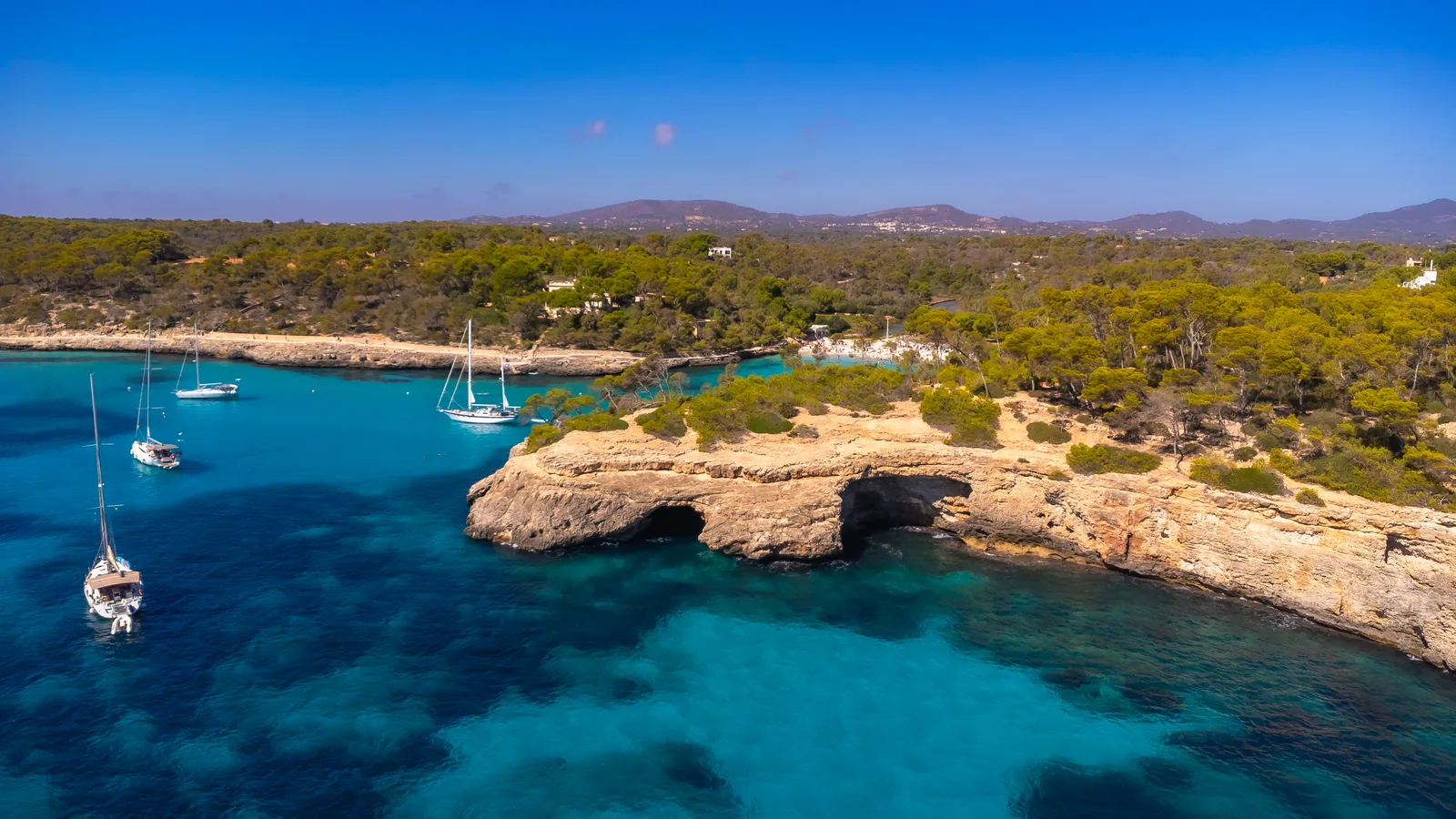 Segelboote, die im türkisfarbenen Wasser vor der Cala Mondrago auf Mallorca vor Anker liegen, dahinter die belebte Bucht und von Kiefern bewachsene Klippen.