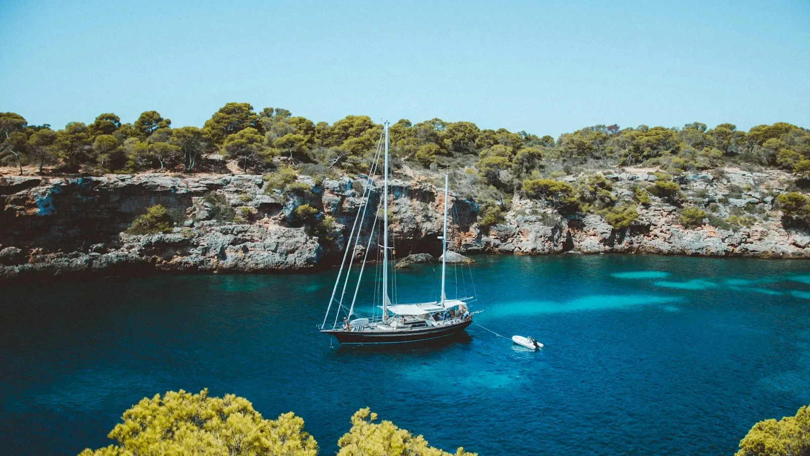 Ein Segelboot, das in einer türkisfarbenen Bucht vor von Kiefern bewachsenen Klippen auf den Balearen vor Anker liegt. Photo by Michael Heuser on Unsplash
